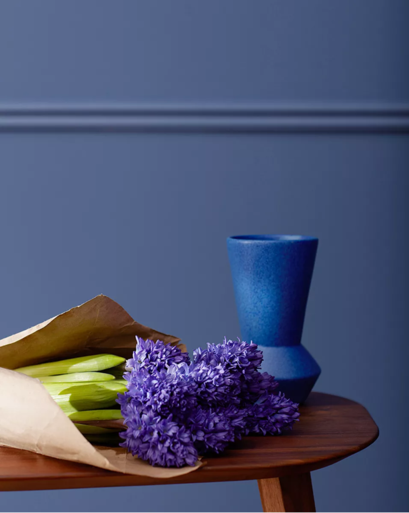 Table holding blue base and purple flowers, positioned in front of wall painted with Benjamin Moore Nova Blue color of the year 